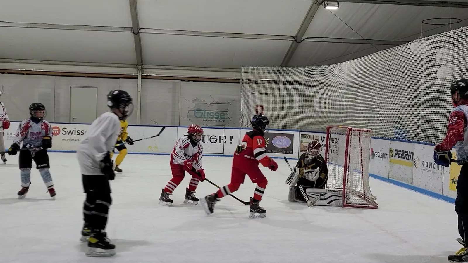 Young hockey players in a game on the mobile rink at Gland under the tent roof, with the goal, boards and sponsor panels around the ice. — Patinoire mobile de Gland