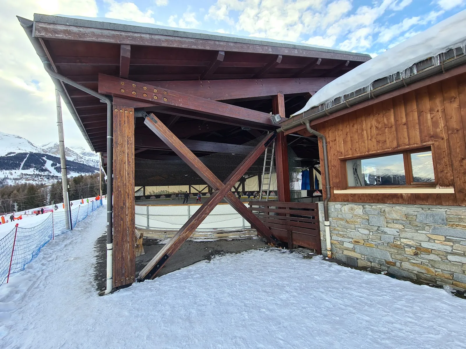 The timber-framed side of the covered rink and its stone-and-wood chalet in the snow, with the ski slopes beyond. — Patinoire Montchavin - Les Coches