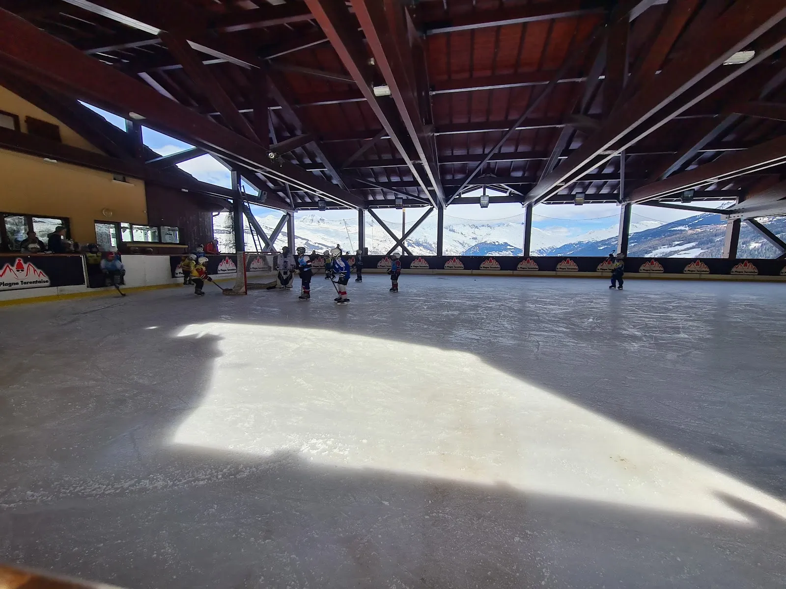 The ice under the timber roof trusses of the open-sided hall, with the mountain view beyond the boards and a few skaters at the far end. — Patinoire Montchavin - Les Coches