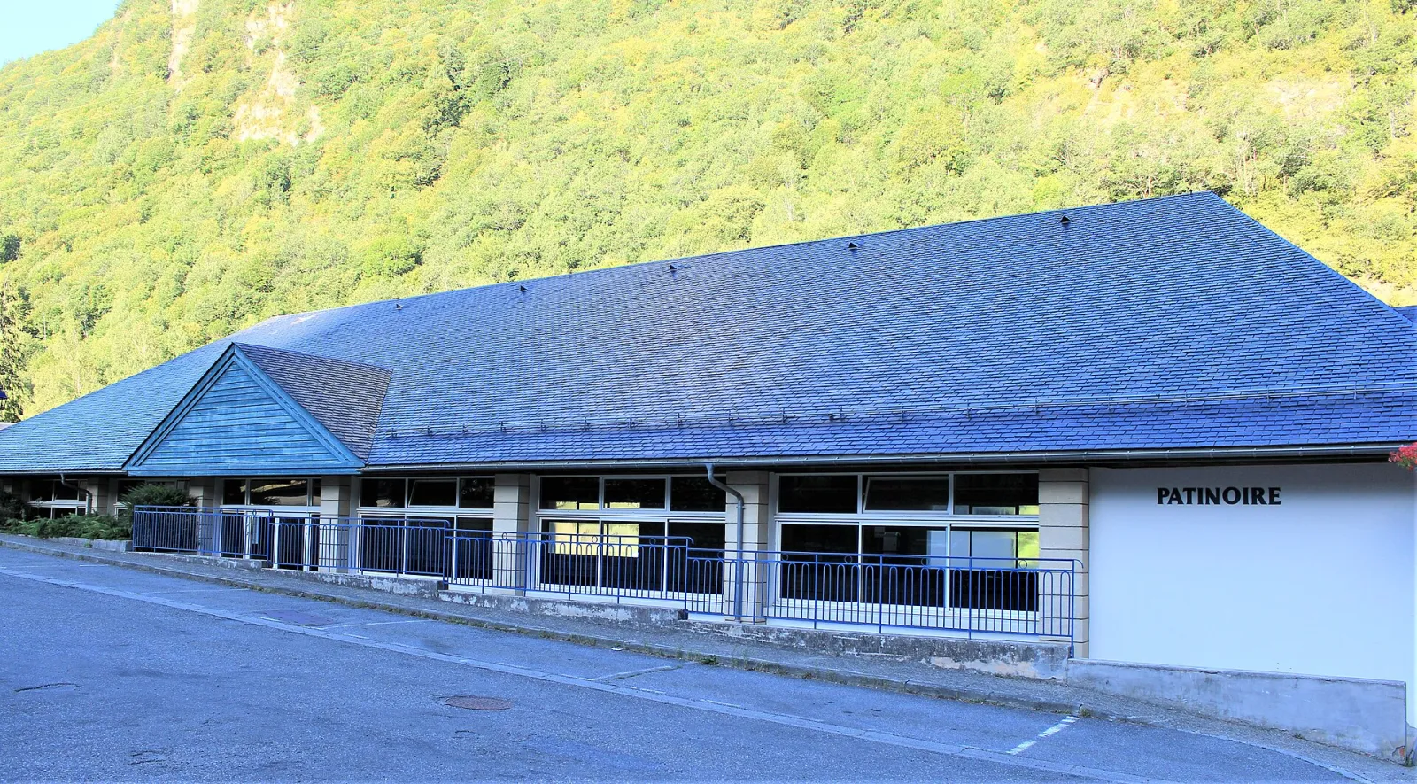 The long low slate-roofed building of the Gèdre patinoire with its glazed front and the word Patinoire on the end wall, under a wooded mountain slope. — Patinoire municipale couverte de Gèdre