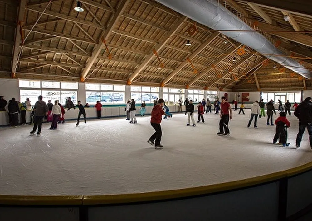 The covered rink at Gèdre under its heavy timber roof, skaters circling the pale ice with large windows along the far side. — Patinoire municipale couverte de Gèdre