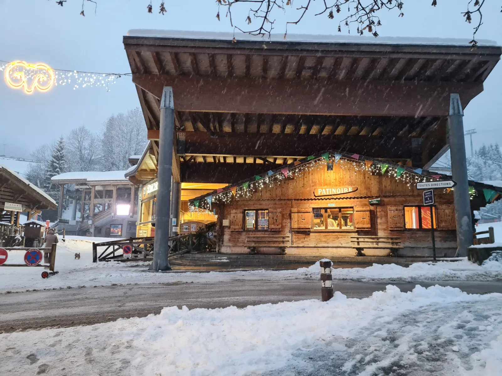 Le bâtiment couvert de la patinoire municipale de La Clusaz un soir de neige, avec son large toit en bois et sa façade de style chalet éclairée. — Patinoire municipale de La Clusaz
