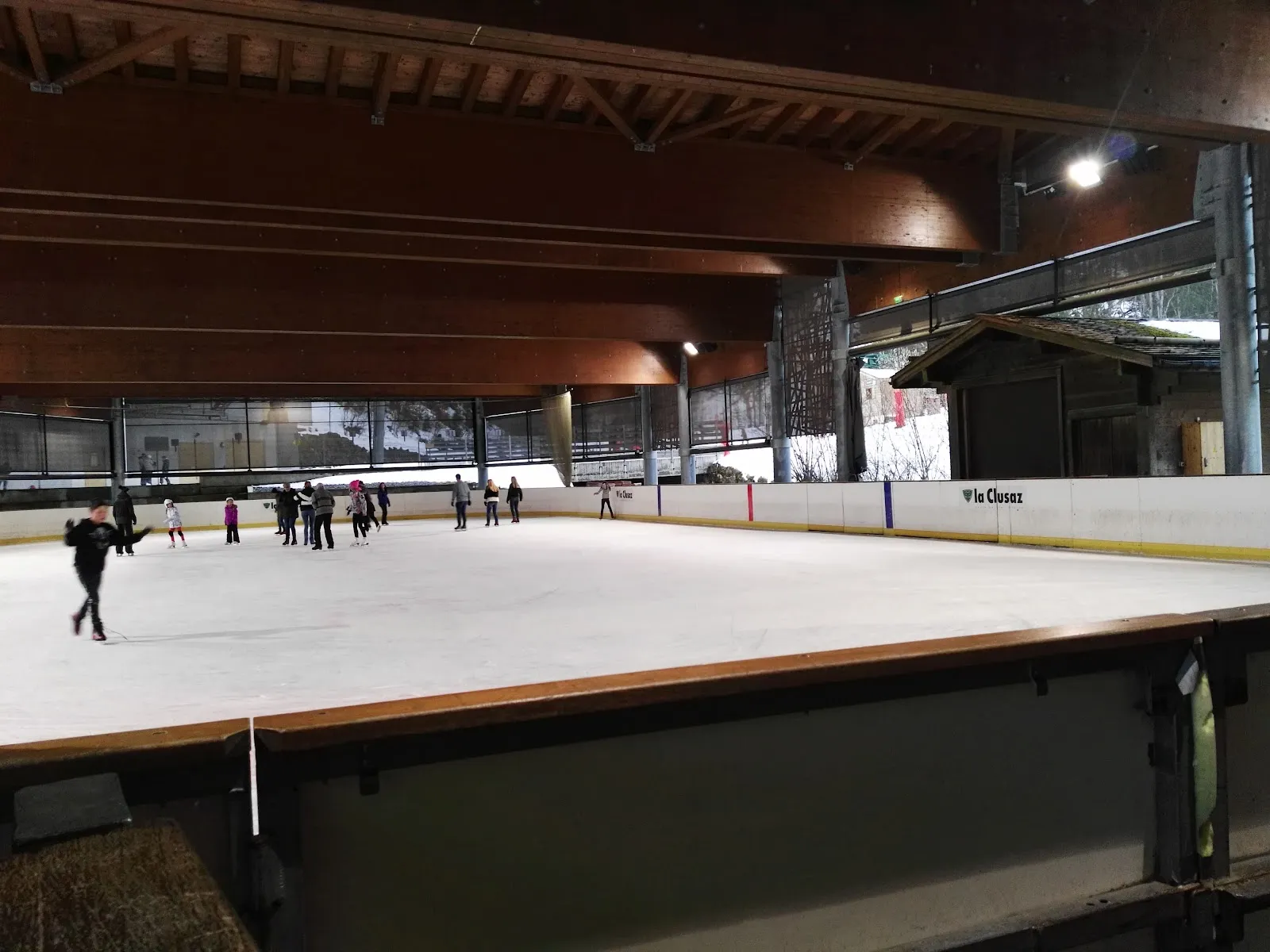 The covered open-air ice under a timber roof, with a few skaters along the boards and the La Clusaz logo on the dasher. — Patinoire municipale de La Clusaz