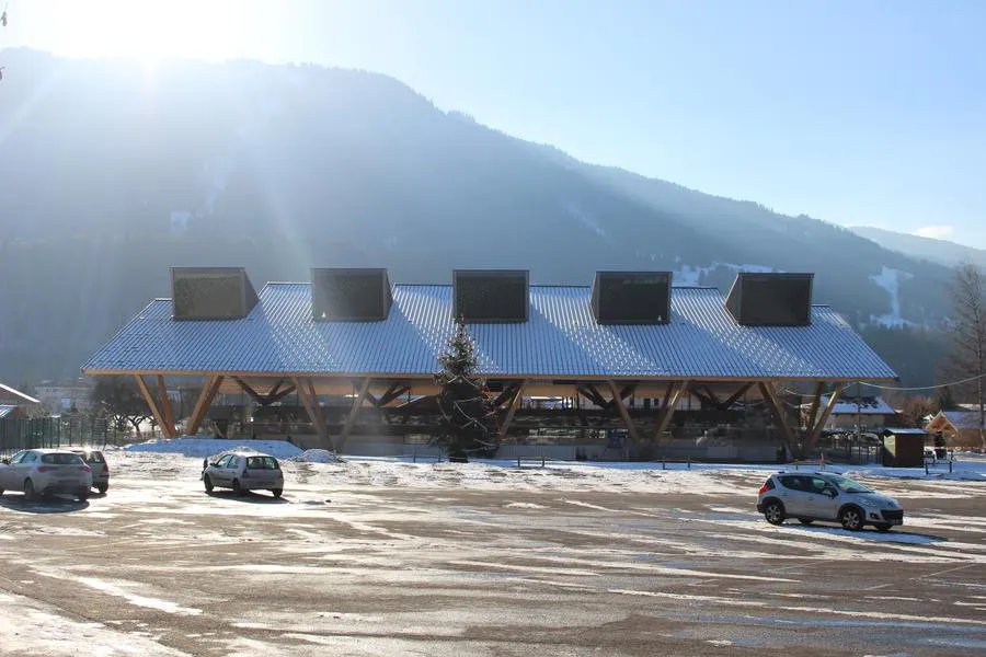 La toiture à ossature bois et le clair-étage de la patinoire couverte de Samoëns vus depuis le parking, avec les montagnes derrière. — Patinoire municipale de Samoëns