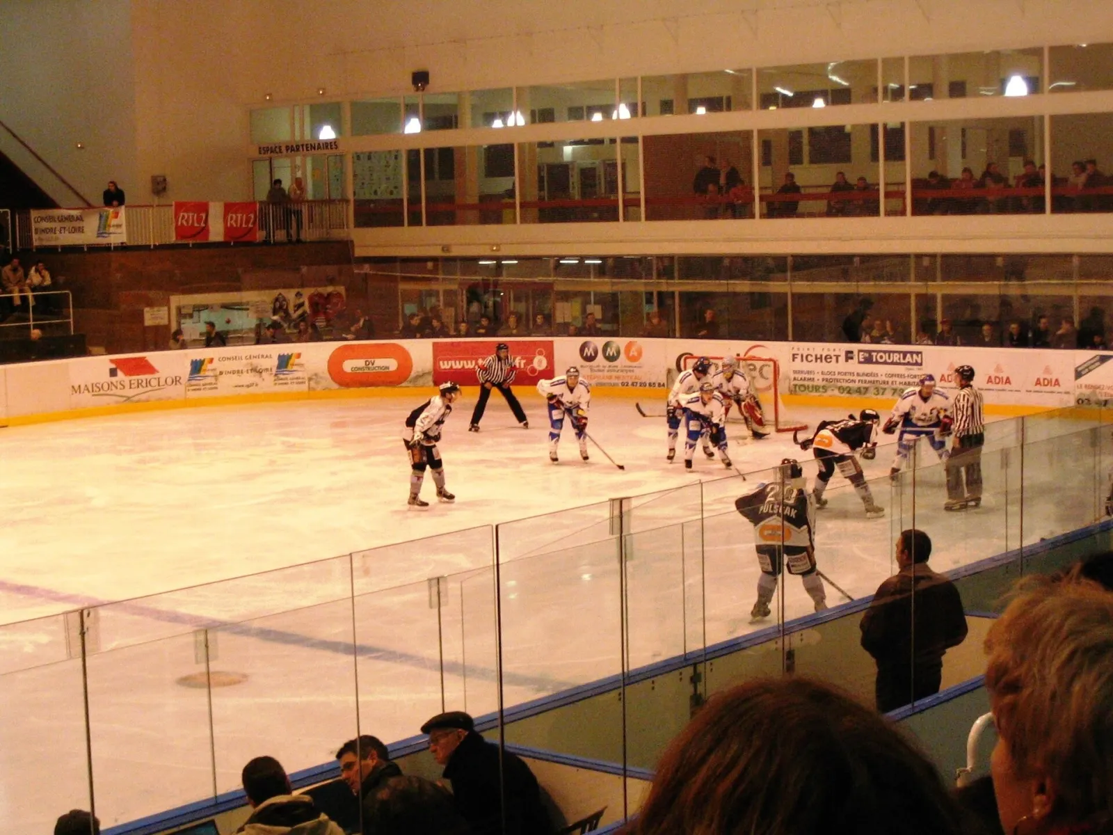 Un match de hockey sur glace en cours à la patinoire de Tours, vu à travers la vitre depuis les gradins, avec des joueurs le long des bandes. — Patinoire municipale de Tours