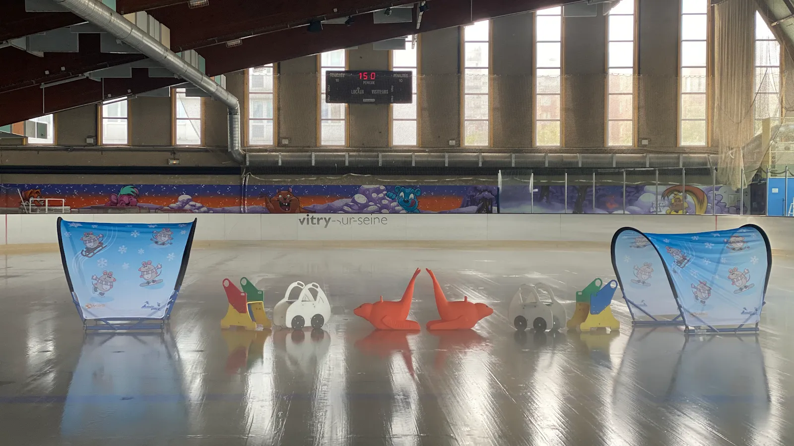 The empty ice with children's skating aids and two pop-up goals in the foreground, the hall's tall windows and the scoreboard behind. — Patinoire municipale de Vitry-sur-Seine