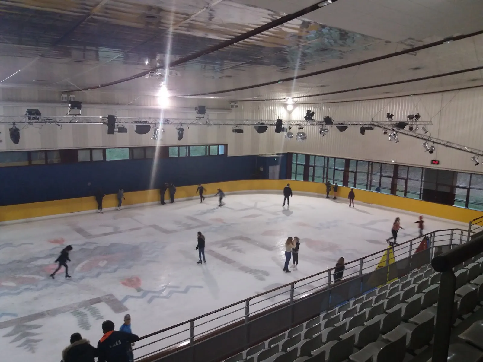 The ice hall seen from the raised seating, with a painted pattern on the ice, a yellow-topped boards line, a glazed wall and a lighting rig overhead. — Patinoire Nautilis