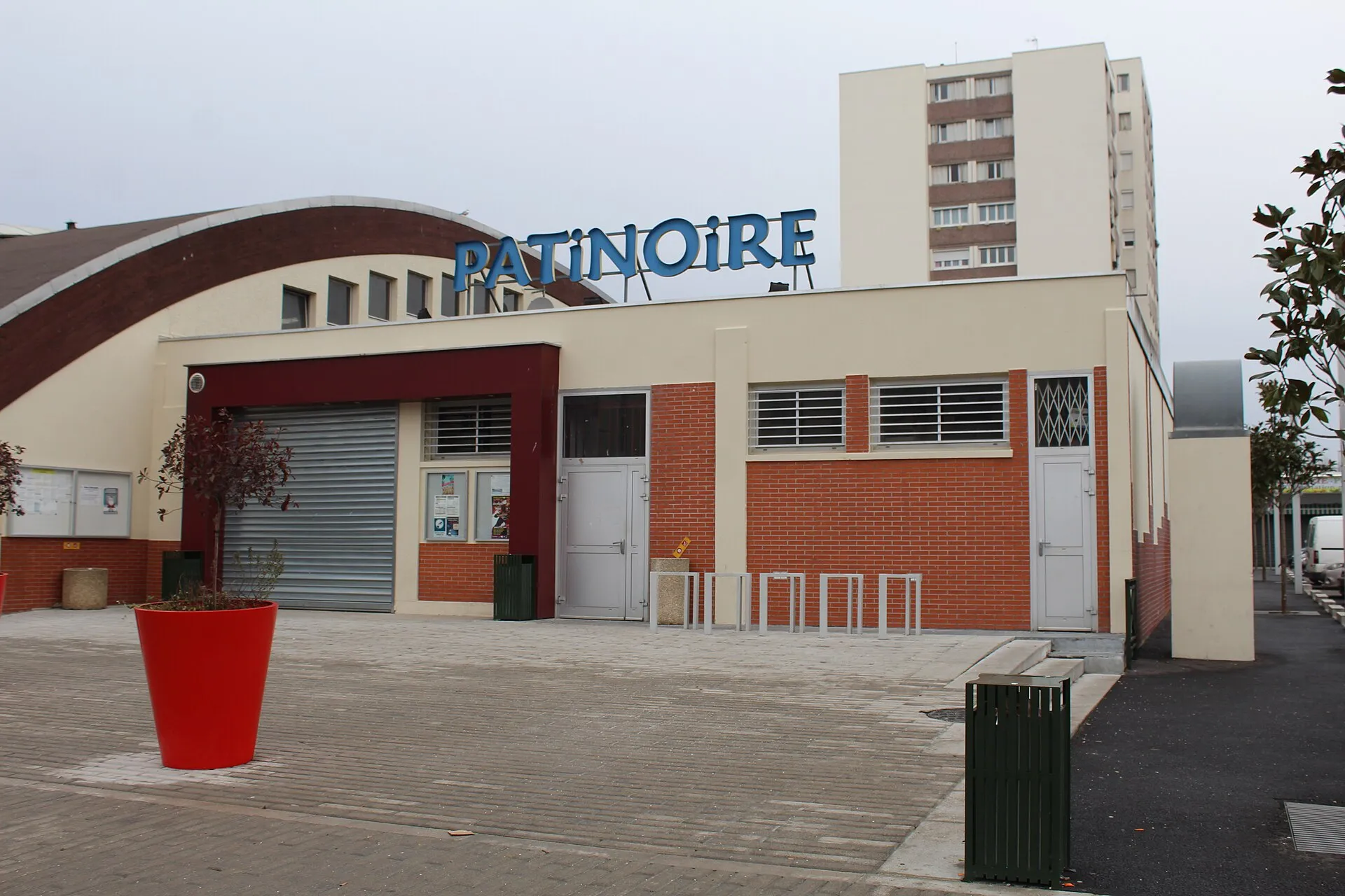 La façade d’entrée de la patinoire de Neuilly-sur-Marne, avec son enseigne bleue PATINOIRE sur la toiture courbe, la façade en brique et enduit, deux jardinières rouges et des râteliers à vélos sur le parvis. — Patinoire municipale de Neuilly-sur-Marne