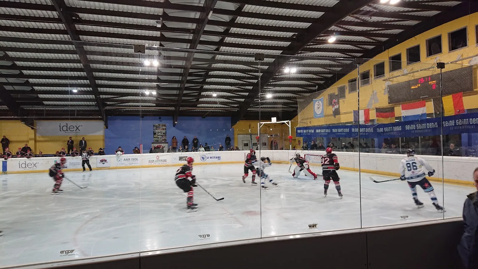 Un match de hockey en cours sur la patinoire de Neuilly-sur-Marne vu à travers la vitre, avec des joueurs sur la glace et des drapeaux internationaux accrochés au mur du fond. — Patinoire municipale de Neuilly-sur-Marne