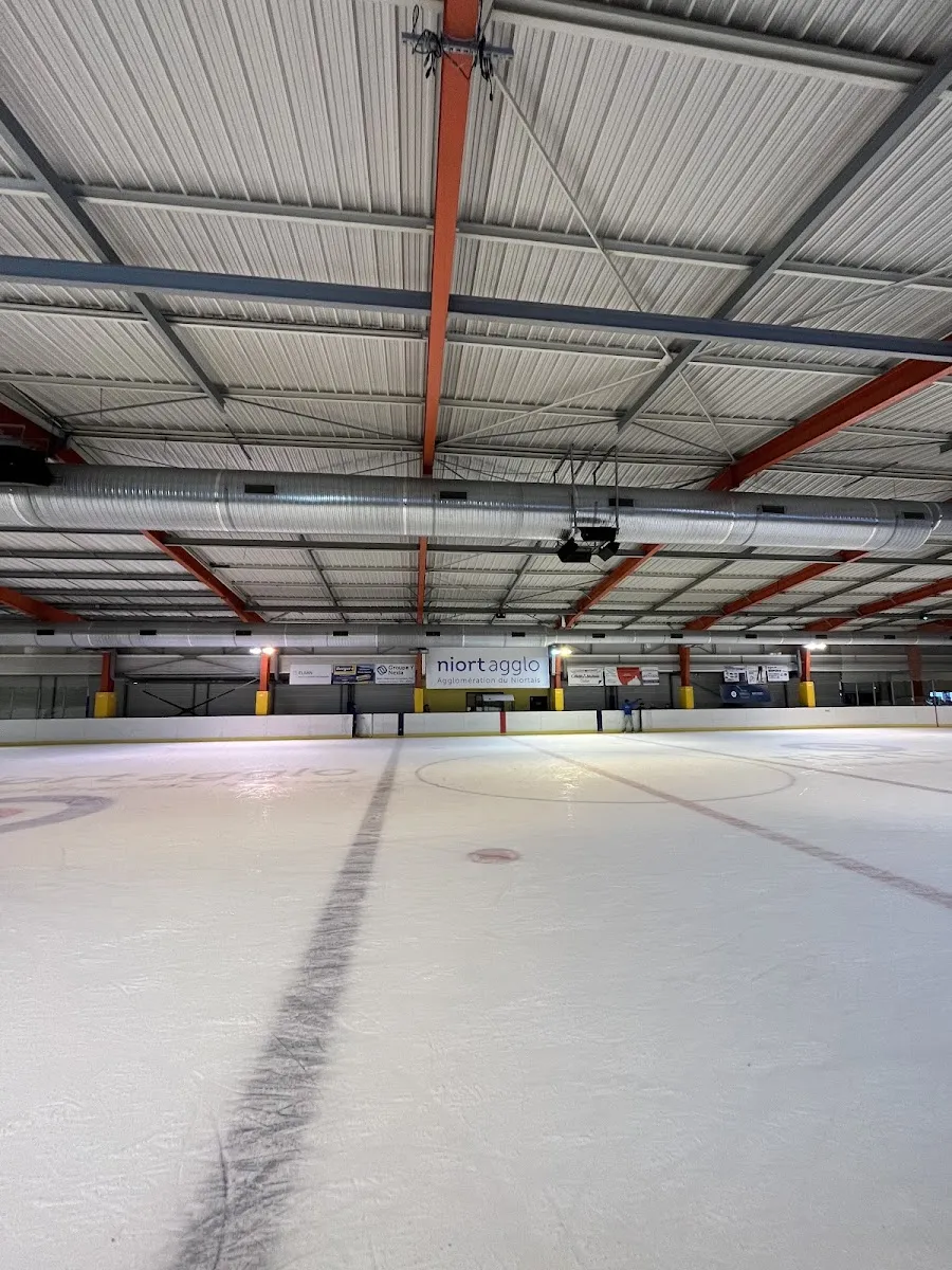 The empty ice of the Niort Agglo rink with the red-and-grey roof structure above the far boards — Patinoire de Niort Agglo