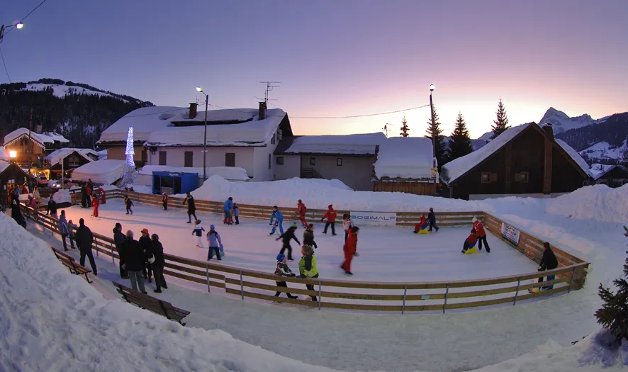 The open-air rink behind its wooden barrier at dusk, among snow-covered village houses with mountains behind. — Patinoire communale de Notre-Dame-de-Bellecombe