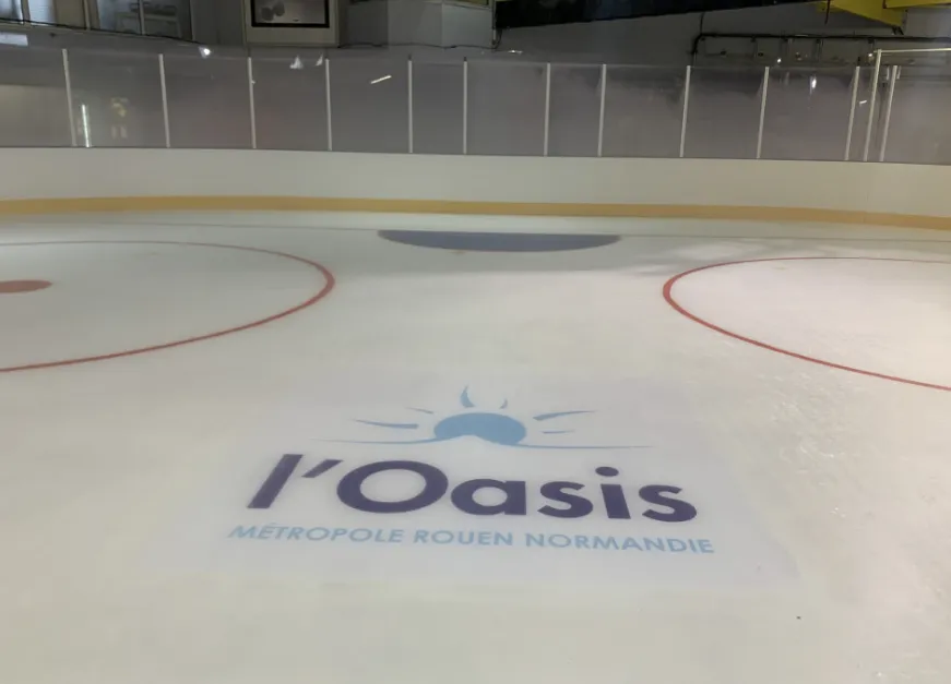 The empty ice with its red lines and faceoff circle and the L'Oasis logo painted on the surface, white boards and glass behind — Patinoire de l'Oasis
