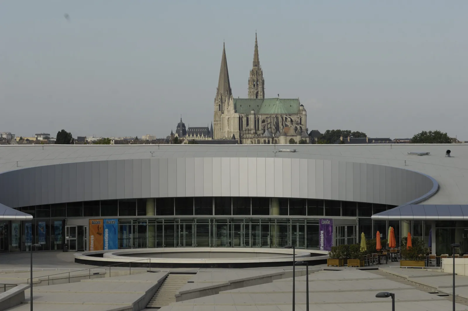 The front of the L'Odyssée complex seen from the forecourt, a low building with a circular silver-clad entrance rotunda and glazed facade, with the Chartres cathedral spires on the skyline. — Patinoire de L'Odyssée