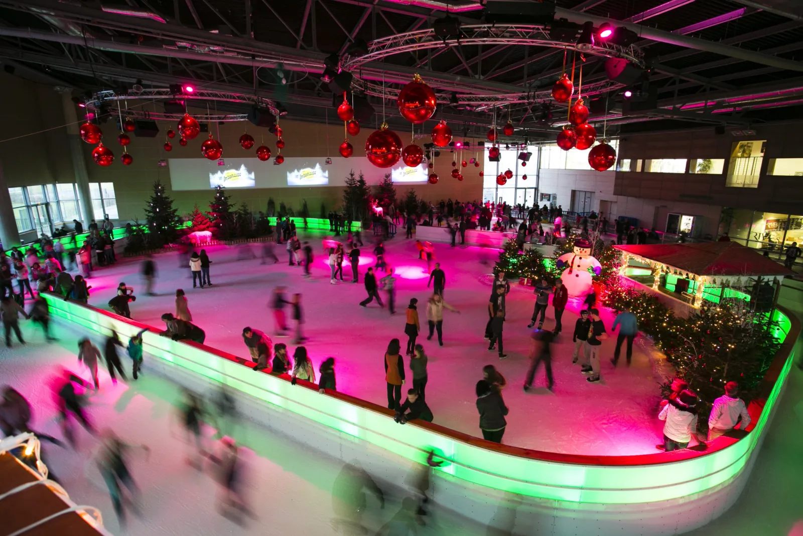 A bird's-eye view of the Odyssée rink during a lit-up Christmas session, skaters circling the ice inside a glowing curved barrier under red baubles. — Patinoire de L'Odyssée