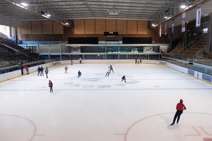The ice and stands of the olympic rink at the Palais des Sports in Megève, with the lighting gantry and wooden-clad end wall above the boards. — Patinoire olympique (Le Palais Megève)