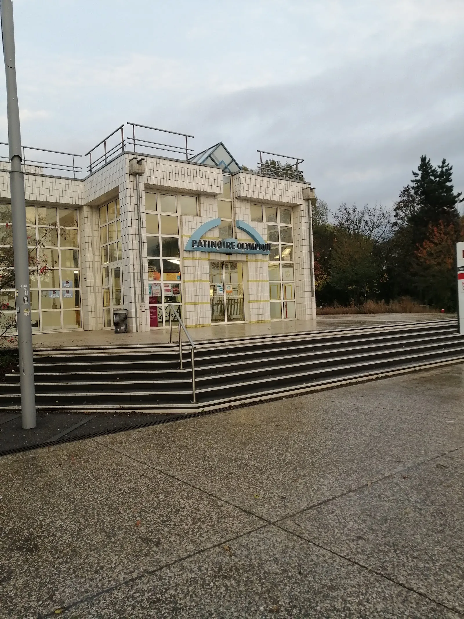 The glazed entrance pavilion of the Patinoire Olympique in Mulhouse, with its name sign above the doors and the wide steps up from the forecourt — Patinoire olympique de Mulhouse