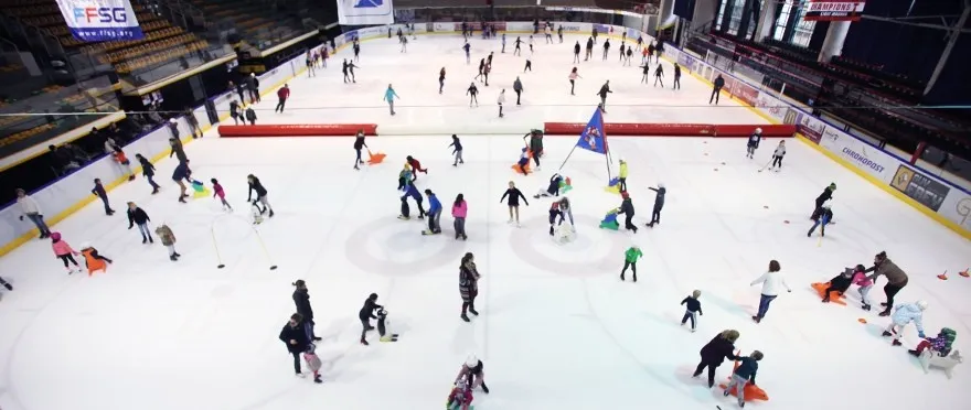 High wide view over the Mulhouse rink ice during a public session, with skaters and children spread across the whole sheet and the boards around the edge. — Patinoire olympique de Mulhouse