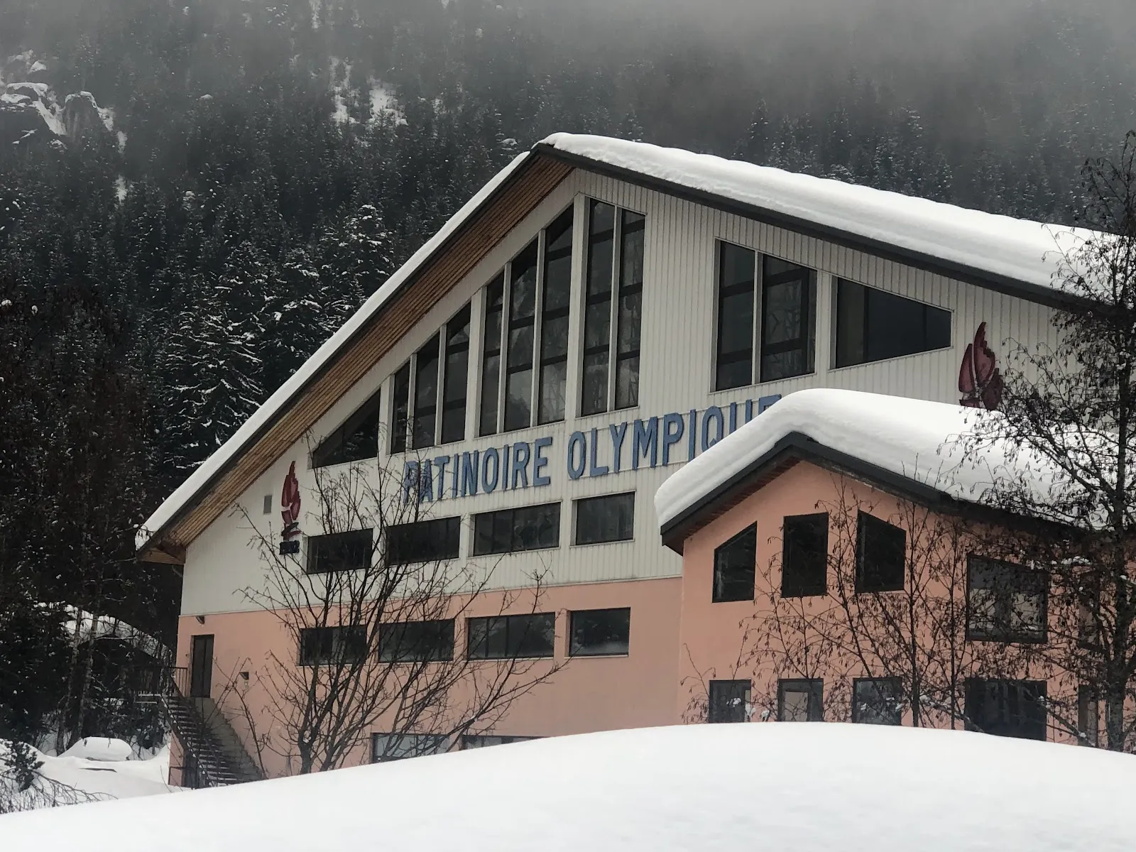 Le bâtiment de la patinoire olympique sous la neige fraîche, avec son toit à deux pans, ses grandes fenêtres et le nom peint sur le pignon, et un versant de montagne couvert de pins derrière. — Patinoire Olympique de Pralognan-la-Vanoise (Le Cristal)