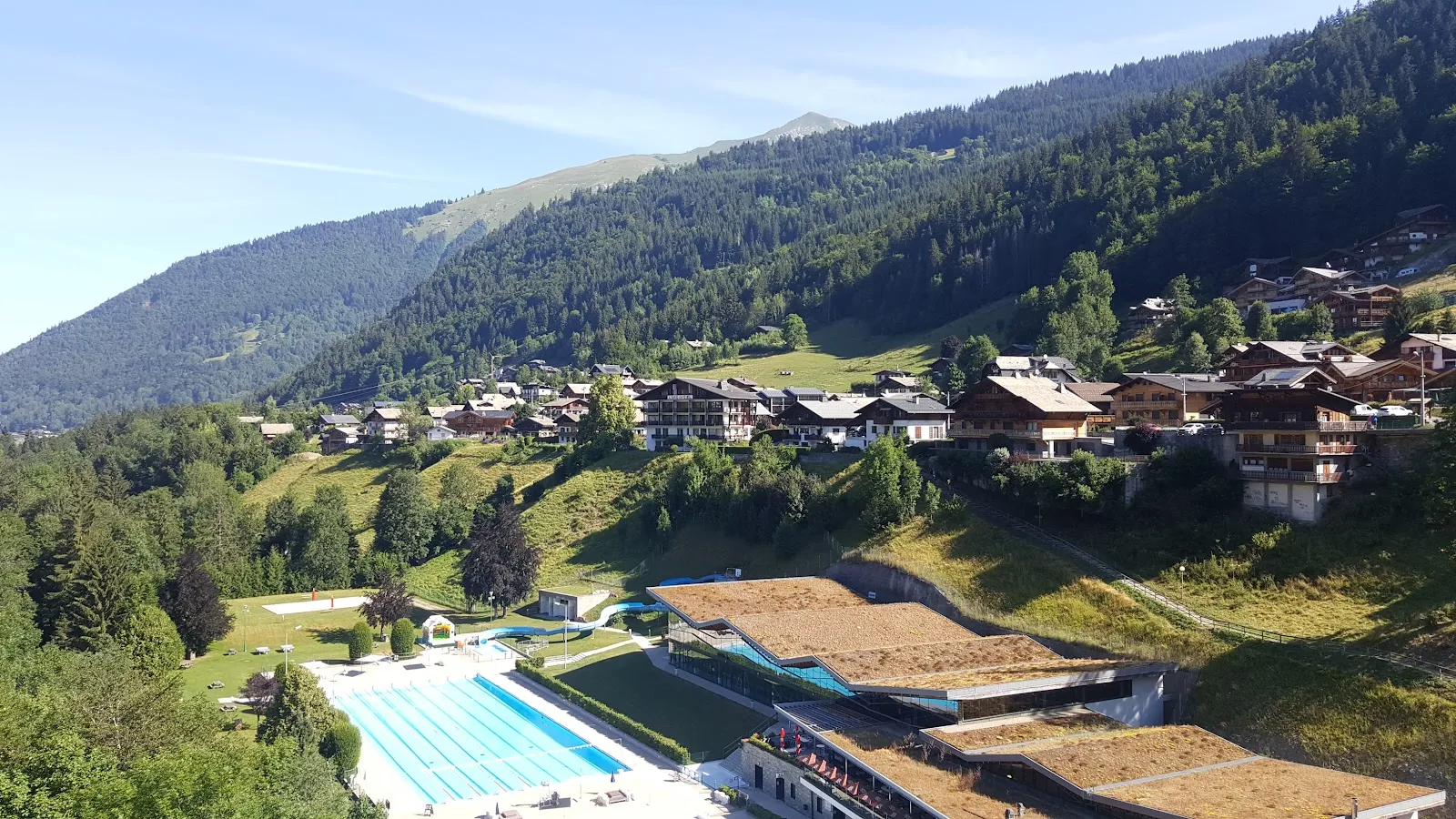 The Palais des Sports in Morzine seen from the hillside, its grass-covered roofs stepping down towards the outdoor swimming pool with chalets and forest behind. — Patinoire du Palais des Sports (Morzine)