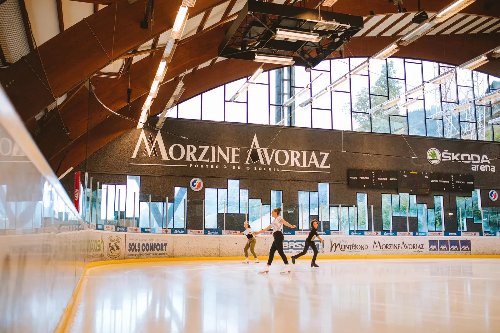 The Morzine Avoriaz ice arena with its curved timber roof and tall windows, a few skaters on the ice and the scoreboard above the far boards. — Patinoire du Palais des Sports (Morzine)
