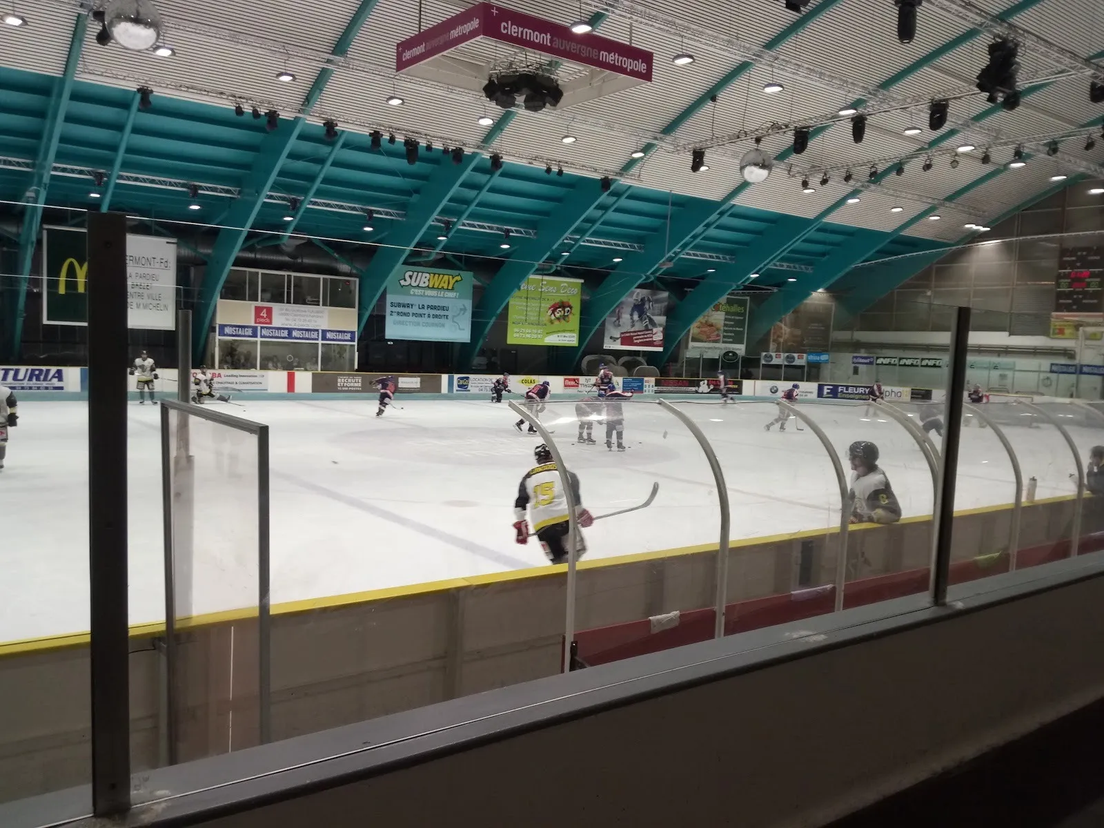 A match on the Clermont-Ferrand rink seen from behind the curved glass, players on the wide ice below the teal-ribbed roof and sponsor boards. — Patinoire Papadakis & Cizeron