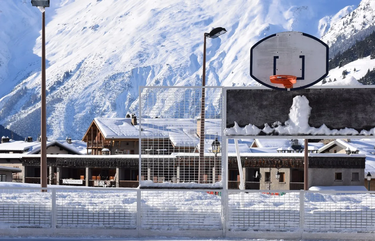 The fenced rink at the Parc de loisirs des Glières in winter, with the village chalets and snowy mountains behind it — Patinoire du Parc de loisirs des Glières