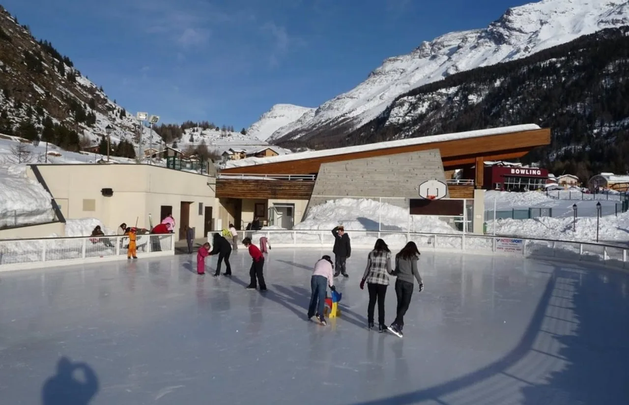 The open-air rink at the Parc de loisirs des Glières in Lanslevillard, with the pavilion and snowy mountains behind and a few skaters on the ice — Patinoire du Parc de loisirs des Glières