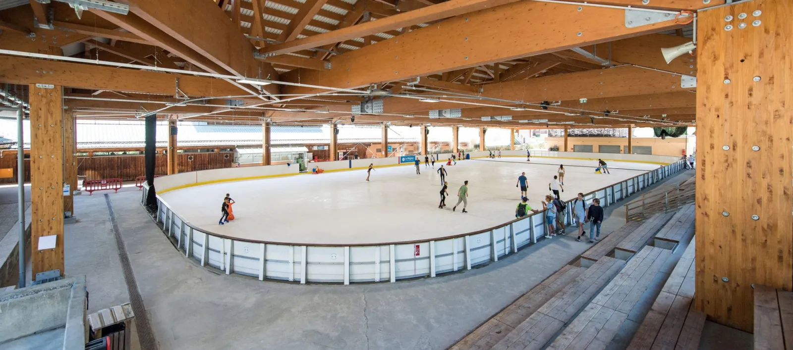 The covered ice of the Valloire rink seen from the stand, with the timber roof posts, the boards and a few skaters far across the ice — Patinoire Philippe Candéloro