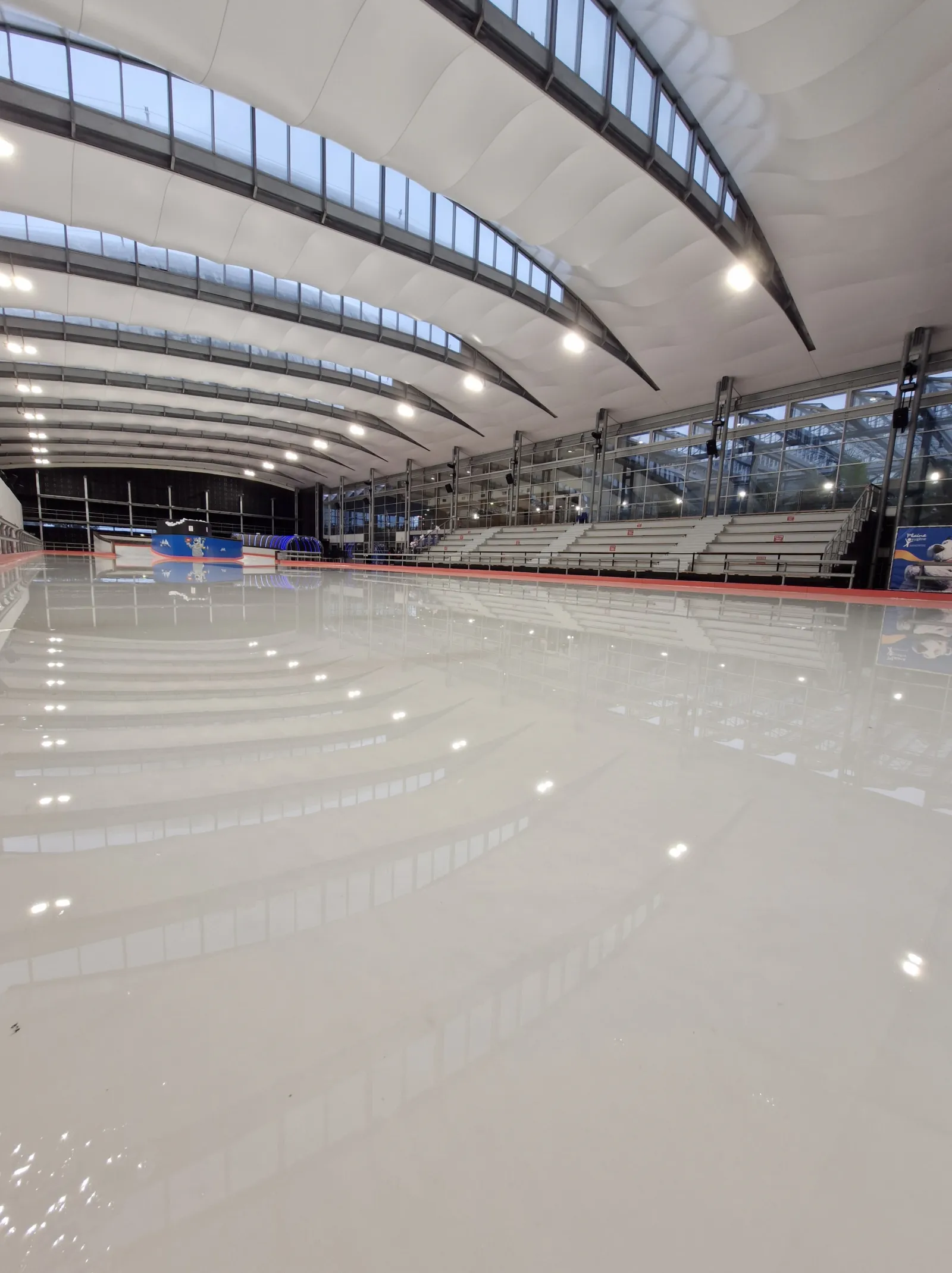 La glace vide vue depuis le bord de la piste, avec une toiture en toile blanche incurvée portant des rangées de puits de lumière au-dessus de la glace et des gradins étagés le long du côté opposé. — Patinoire de la Plaine Oxygène