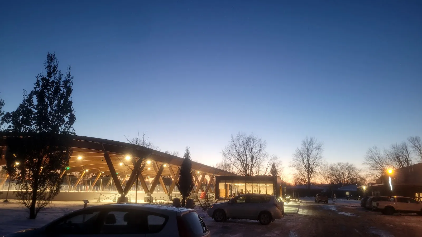 The covered refrigerated rink in Dorval at dusk, under its timber roof, with the lit pavilion beside it — Patinoire réfrigérée couverte de Dorval
