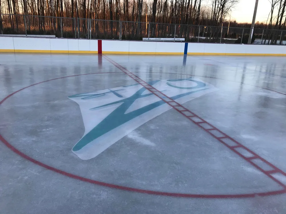 The centre-ice logo and red circle on the refrigerated rink at parc Quatre-Saisons, with white boards, a lit fence line and bare trees behind. — Patinoire réfrigérée du parc Quatre-Saisons (Saint-Zotique)