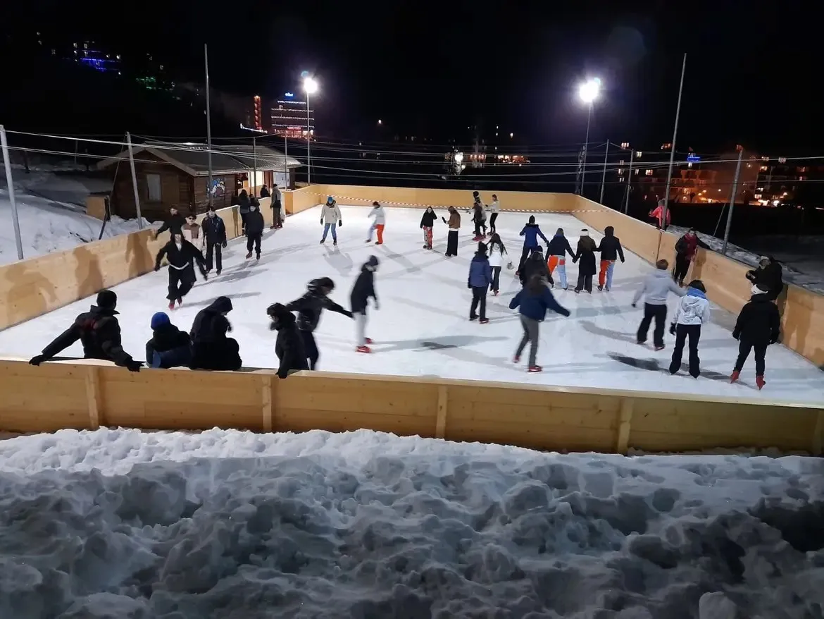 La patinoire en plein air de Risoul la nuit, des patineurs sur la glace enneigée à l’intérieur d’une barrière en bois, avec un chalet éclairé derrière. — Patinoire de Risoul