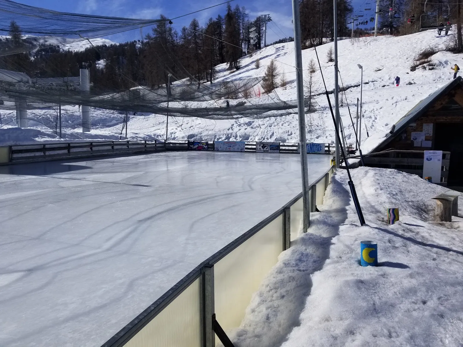 La patinoire extérieure vide de Risoul 1850 vue depuis le bord, avec le talus de neige, le filet au-dessus de la pente et le chalet à côté de la glace — Patinoire de Risoul