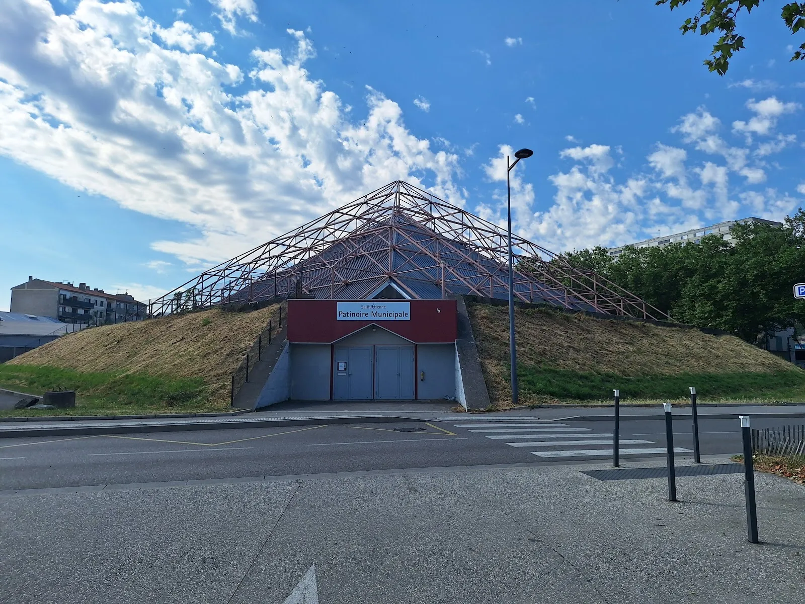 Le toit en forme de pyramide de la patinoire de Saint-Étienne qui s’élève au-dessus d’un talus herbeux, avec l’enseigne rouge « Patinoire Municipale » au pied de l’entrée. — Patinoire de Saint-Étienne