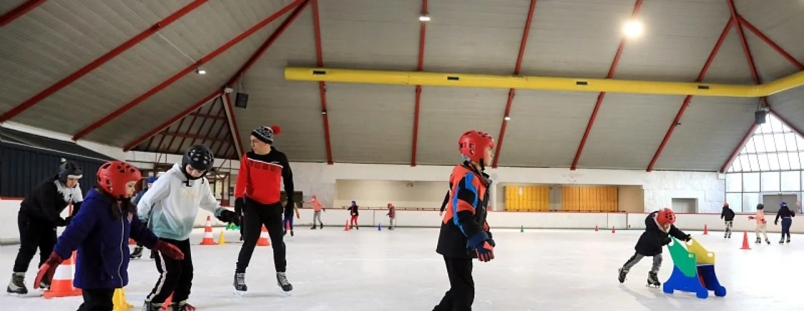 De jeunes patineurs casqués s’entraînant avec des cônes sur la glace de la patinoire de Saint-Étienne, sous son toit pyramidal à nervures rouges. — Patinoire de Saint-Étienne