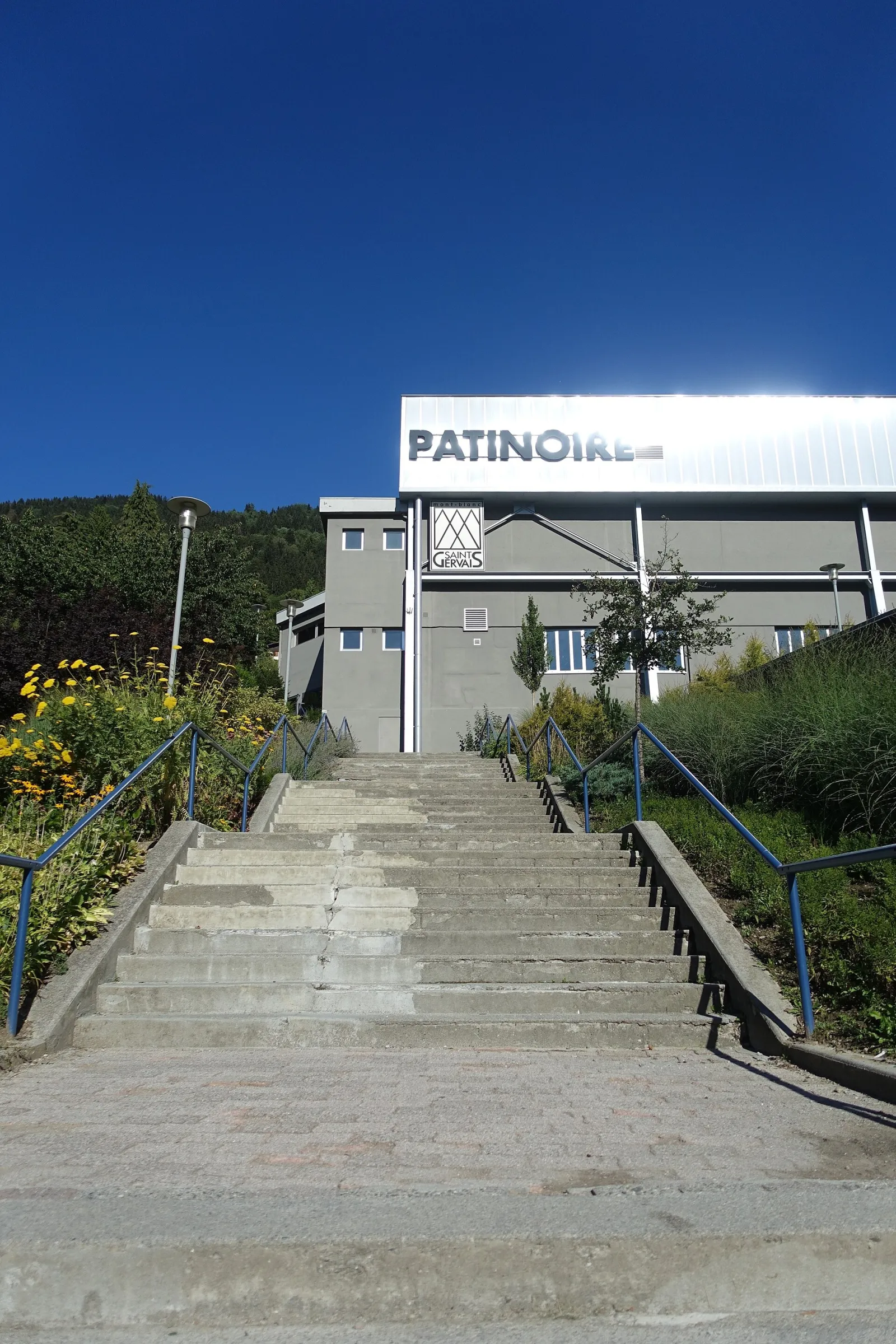 Le haut de la façade du bâtiment de la patinoire de Saint-Gervais, avec le mot PATINOIRE sur sa ligne de toit blanche contre un ciel bleu, et une pente boisée derrière. — Patinoire municipale de Saint-Gervais