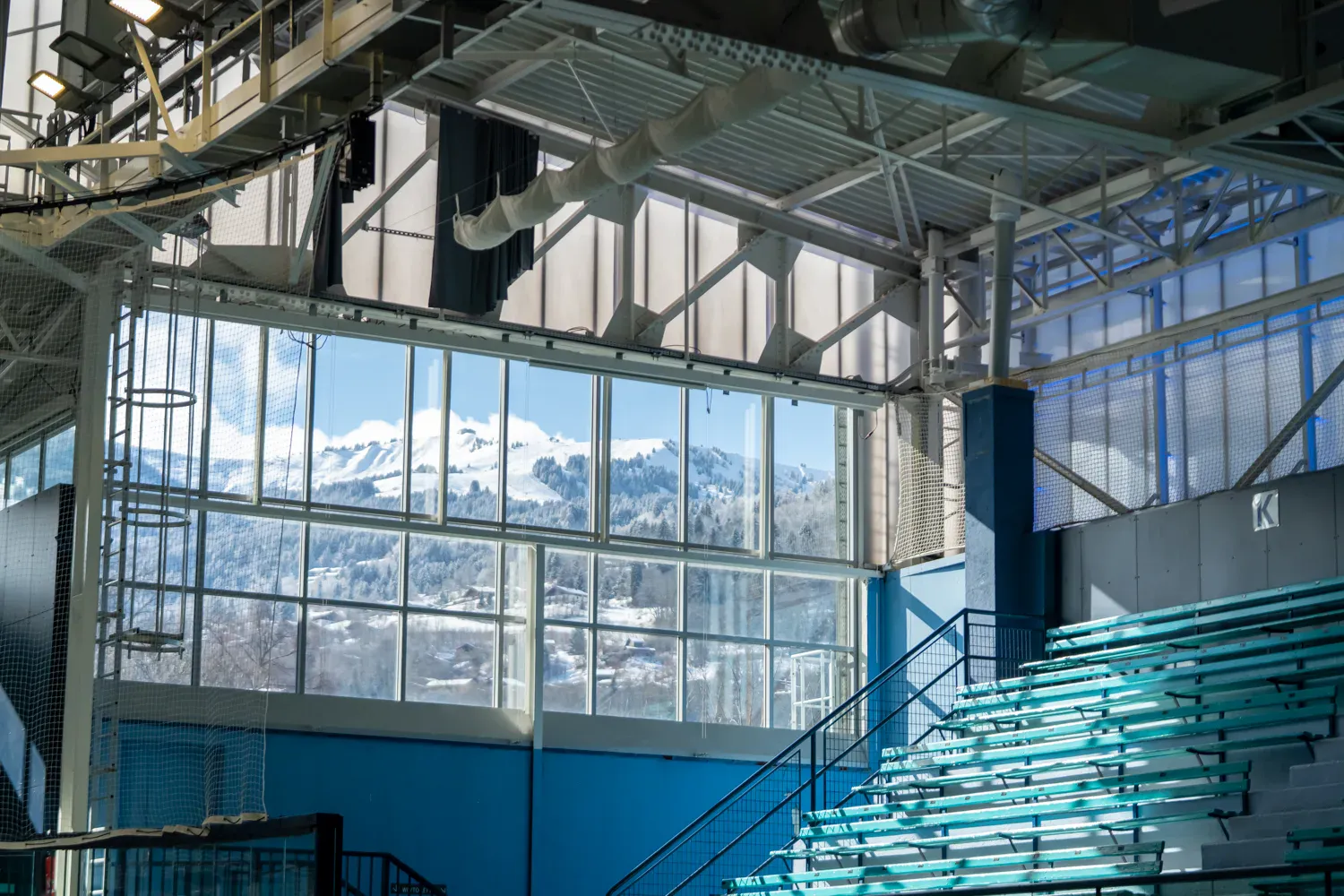 La charpente en acier et les gradins bleu-vert de la patinoire de Saint-Gervais, avec un mur de baies vitrées donnant sur les montagnes. — Patinoire municipale de Saint-Gervais