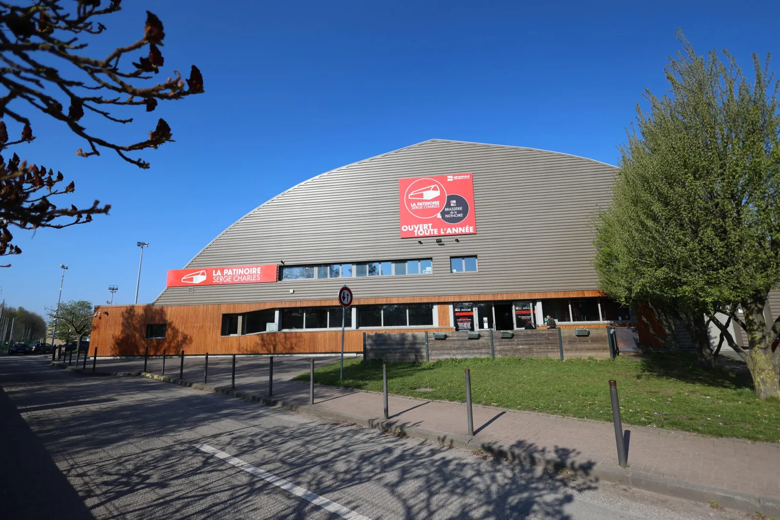 The arched metal-clad building seen from the street in clear light, with a red rink sign on the facade, a red La Patinoire Serge Charles banner and a timber-clad low wing. — Patinoire Serge Charles