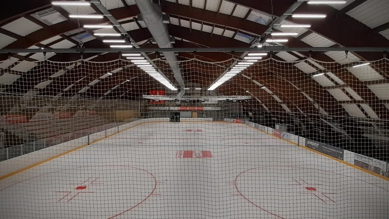 The empty Wasquehal ice under its curved timber vault, with the red face-off circles marked on the white surface and the stands along the sides. — Patinoire Serge Charles