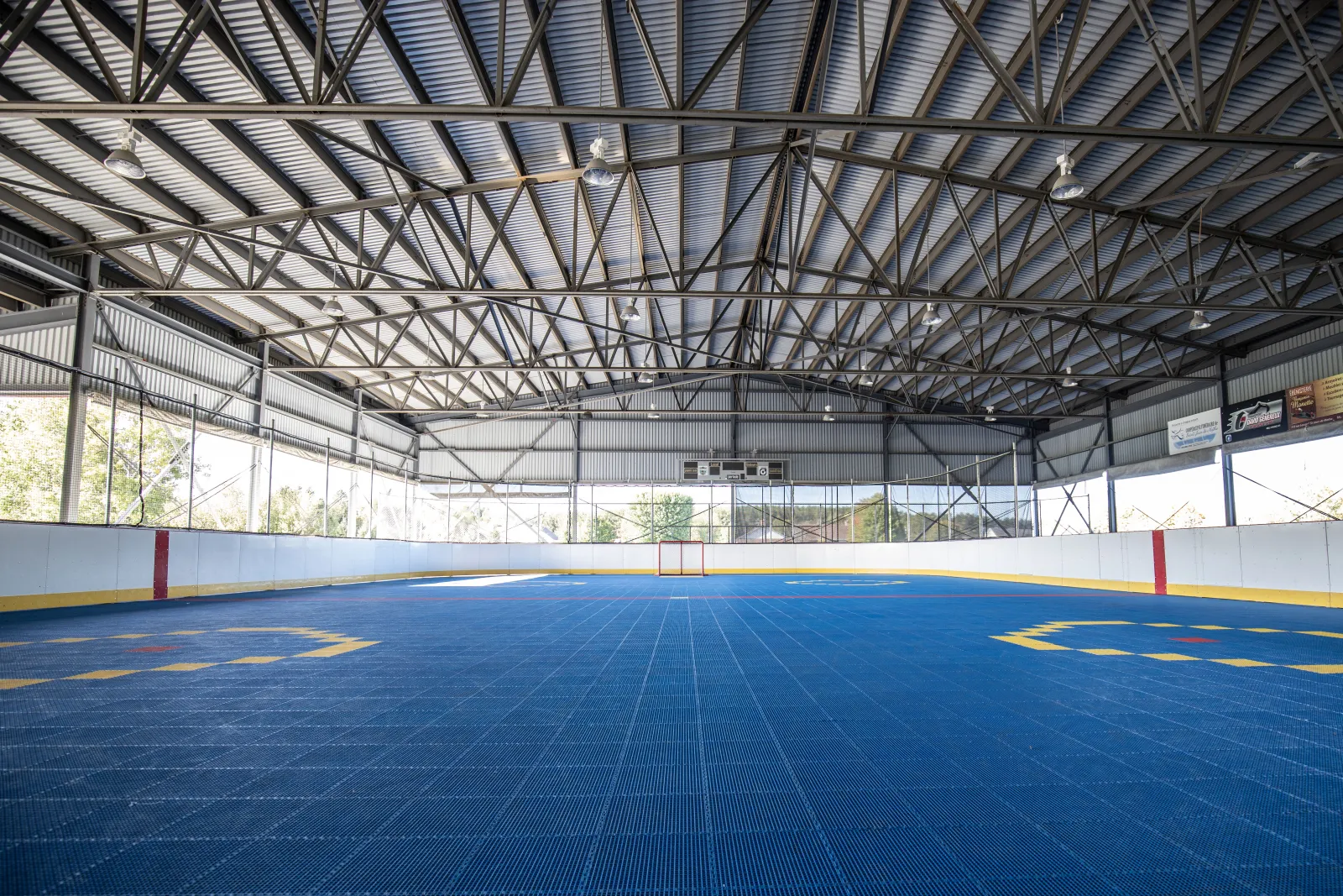 Dans la patinoire couverte du parc Donat-Gadoury en été : le plancher bleu du terrain sportif sous la charpente d'acier du toit, entouré des bandes. — Patinoire sous le préau du parc Donat-Gadoury (Saint-Jean-de-Matha)