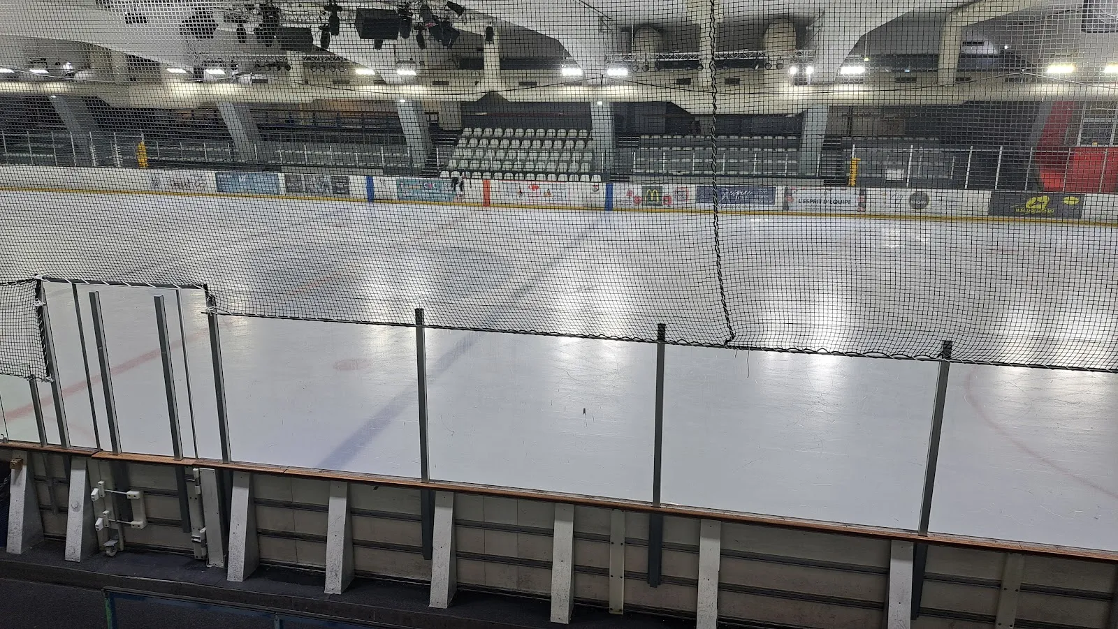 La glace vide de la patinoire municipale de Courbevoie vue des gradins, derrière le filet de protection, avec les panneaux de sponsors et les places assises couvertes de l’autre côté — Patinoire municipale Thierry Monier