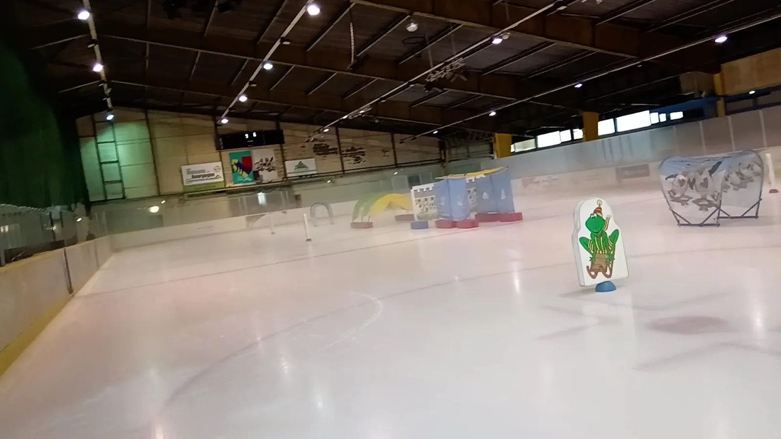 The ice along the long side of the rink with children's teaching aids and training props near the boards, under the hall's dark timber roof. — Patinoire Trimolet