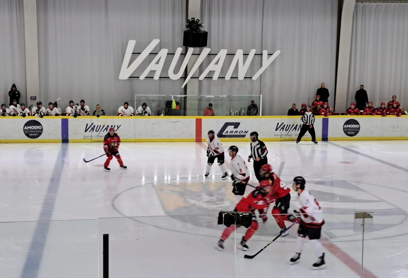 Un match de hockey sur glace : des joueurs en rouge et blanc près de la bande, un banc le long de la vitre au fond et le lettrage Vaujany sur le mur du fond, au-dessus de la bande. — Patinoire de Vaujany