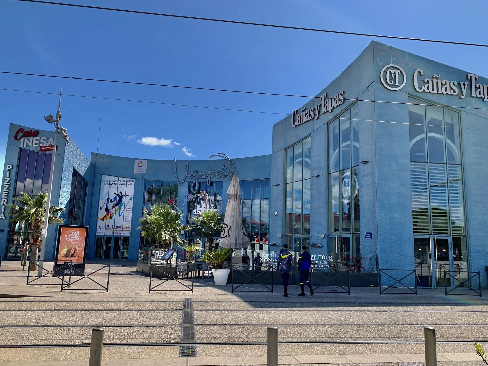 L’entrée de Végapolis dans le complexe Odysseum : une façade bleue incurvée avec l’enseigne « Vegapolis » en écriture cursive au-dessus de portes vitrées, une banderole de championnat de patinage artistique et des terrasses de restaurants de part et d’autre. — Patinoire Végapolis