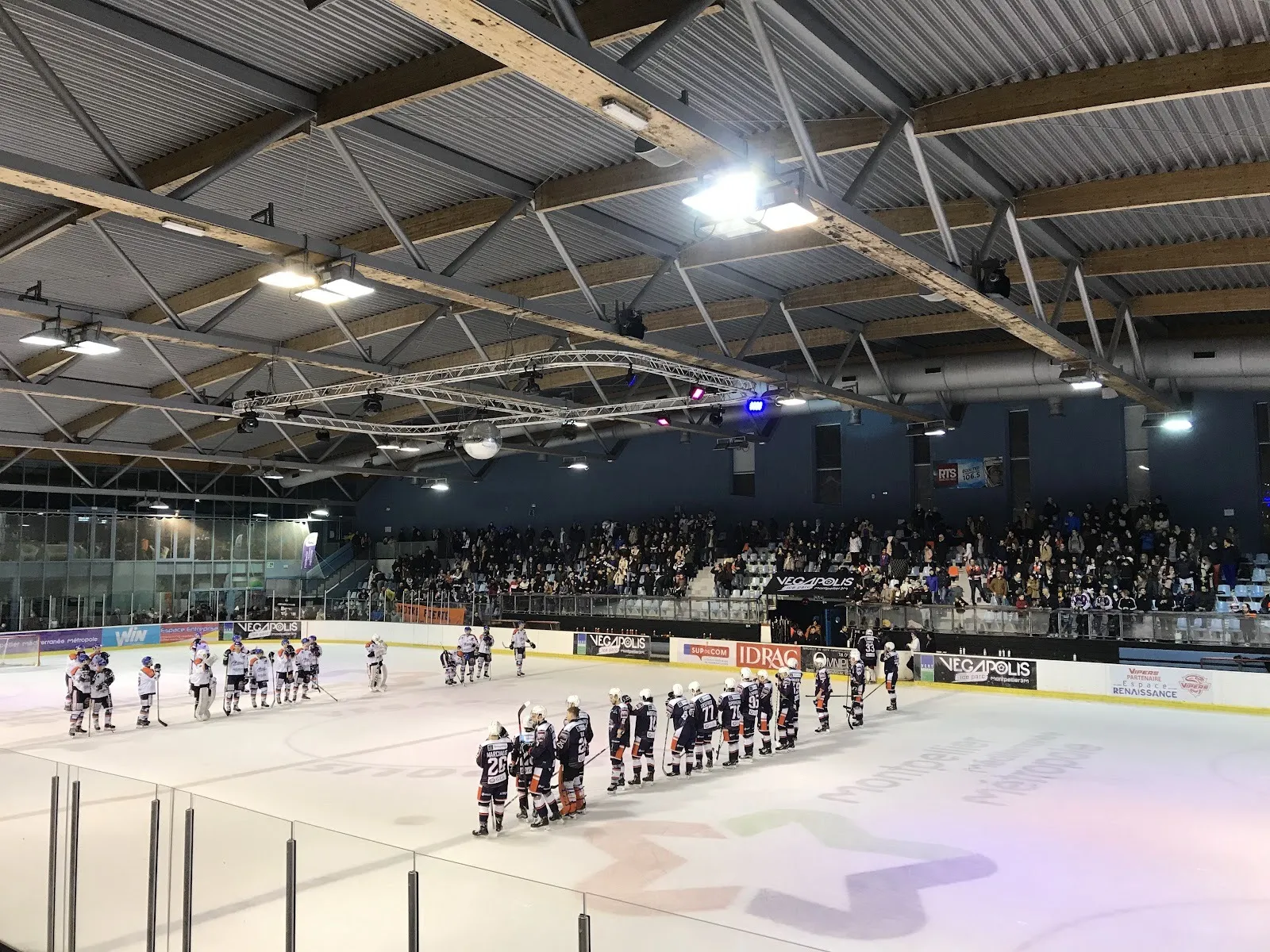 Des joueurs alignés sur la glace de la patinoire Végapolis après un match, avec les gradins bondés et les panneaux de sponsors derrière eux sous le toit d’acier. — Patinoire Végapolis