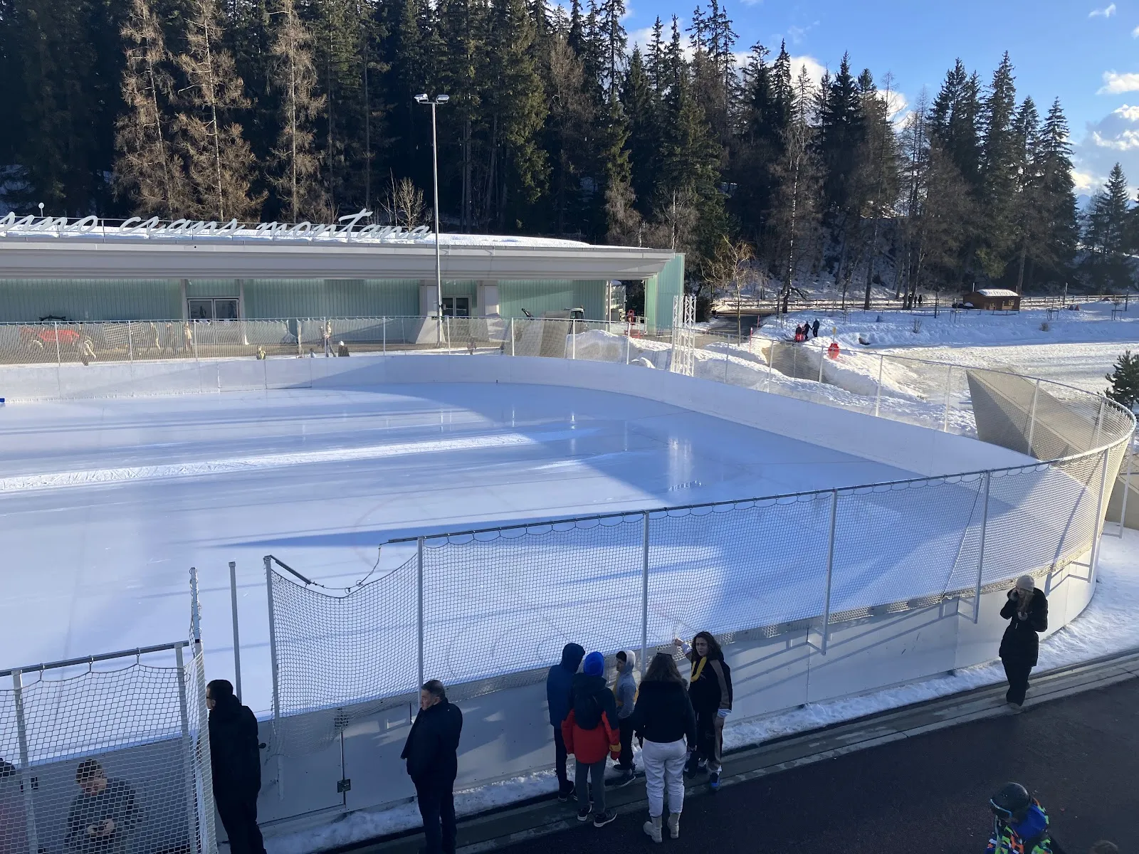 The outdoor Ycoor rink in Crans-Montana seen from above with its curved boards, the glass building behind and skaters on the ice — Patinoire d'Ycoor (Crans-Montana)