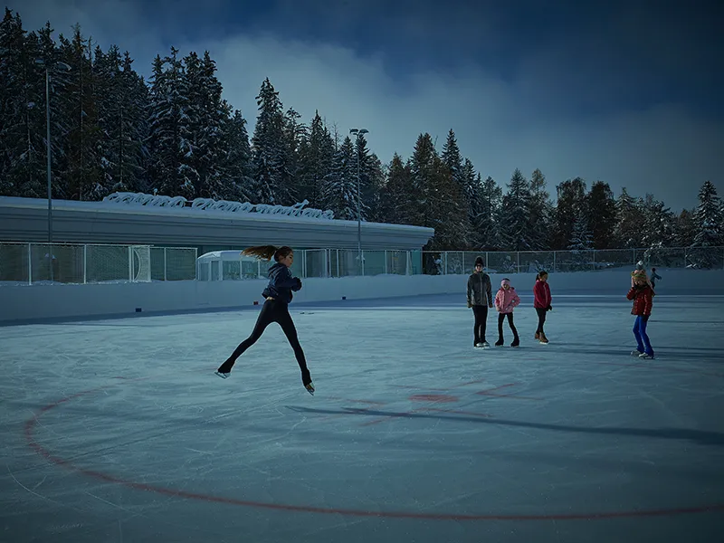 The open-air Ycoor rink at dusk: a skater leaps in the foreground and a few skaters stand and glide in the distance on the blue-white ice, with the rink's glass-topped boards, a low building and snowy fir forest behind. — Patinoire d'Ycoor (Crans-Montana)