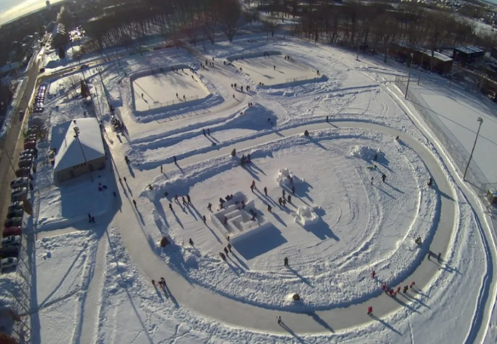 An aerial view of the rinks and the skating loop at Parc Olympia in winter, with skaters small on the ice and the chalet beside them. — Patinoires du parc Olympia (Deux-Montagnes)