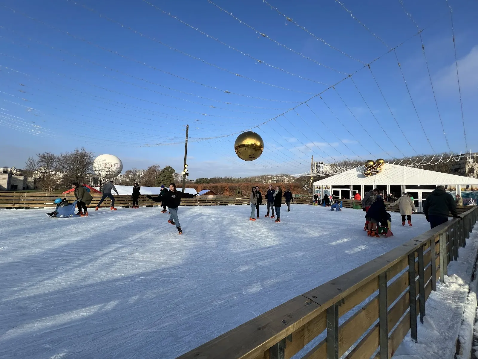 Skaters on the daytime ice of Patins en Folie in Parc André-Citroën, with the park balloon and the light canopy above the boards — Patins en Folie au Parc André-Citroën
