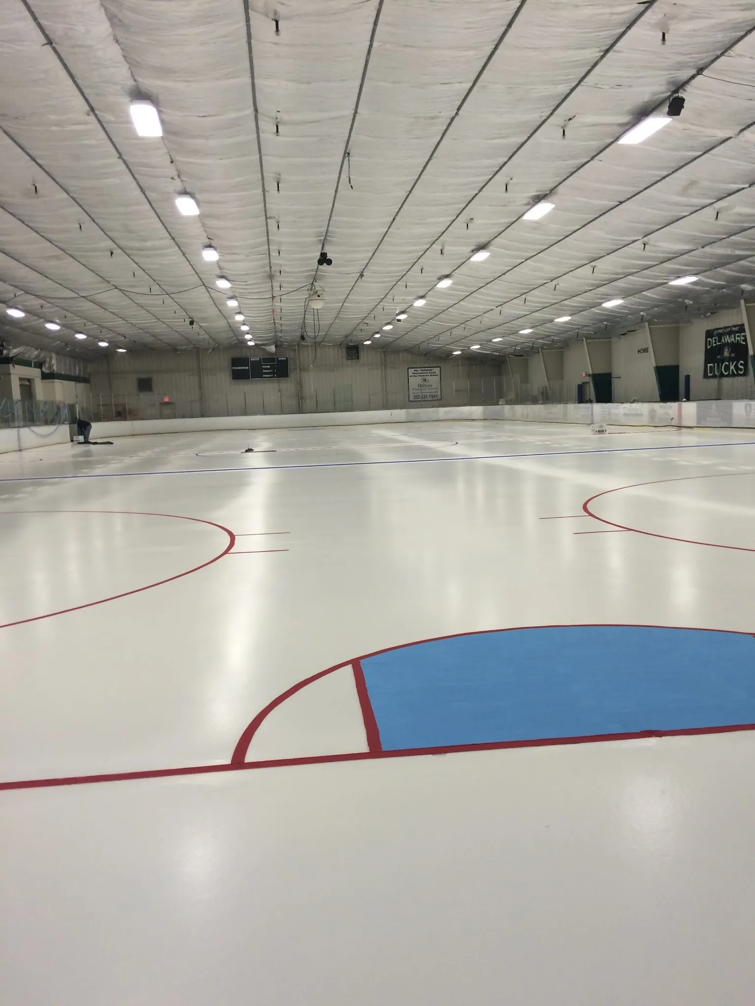 An empty sheet seen from the end, a red line and blue crease on the white ice under a long ceiling of lights. — The Patriot Ice Center